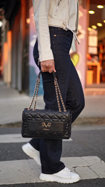 A person walking on a city street carrying a black quilted leather shoulder bag with gold tone chain straps and a gold logo on the front. The person is wearing a cream jacket, dark blue jeans, and white sneakers. 19V69 Italia, Monza Outlet, Monza Fashion, Shein Outlet