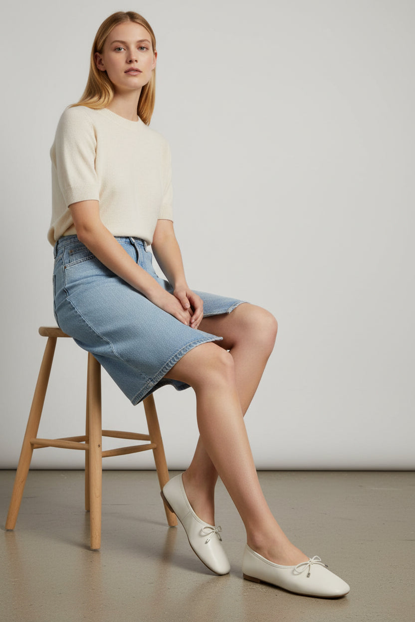 Woman sitting on a wooden stool wearing a light-colored top, denim skirt, and white shoes against a plain background. Aldo, Monza Outlet, Monza Fashion, Shein Outlet