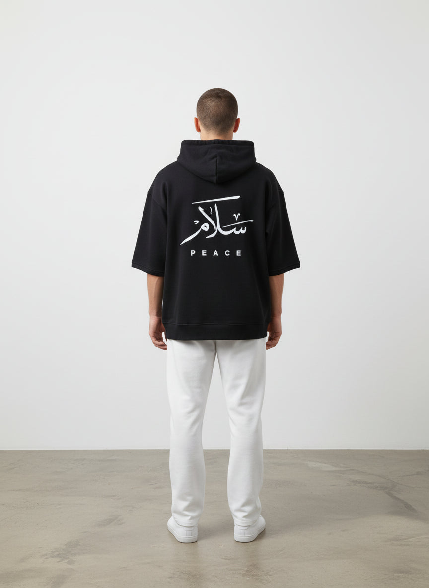 Person wearing a black hoodie with Arabic text and 'PEACE' on the back, standing against a white wall. Antler, Monza Outlet, Monza Fashion, Shein Outlet