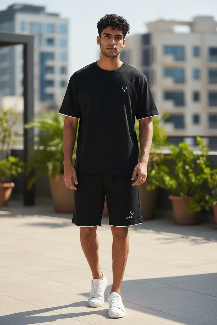 Man wearing a black t-shirt and shorts standing on a rooftop with plants and buildings in the background. Antler, Monza Outlet, Monza Fashion, Shein Outlet
