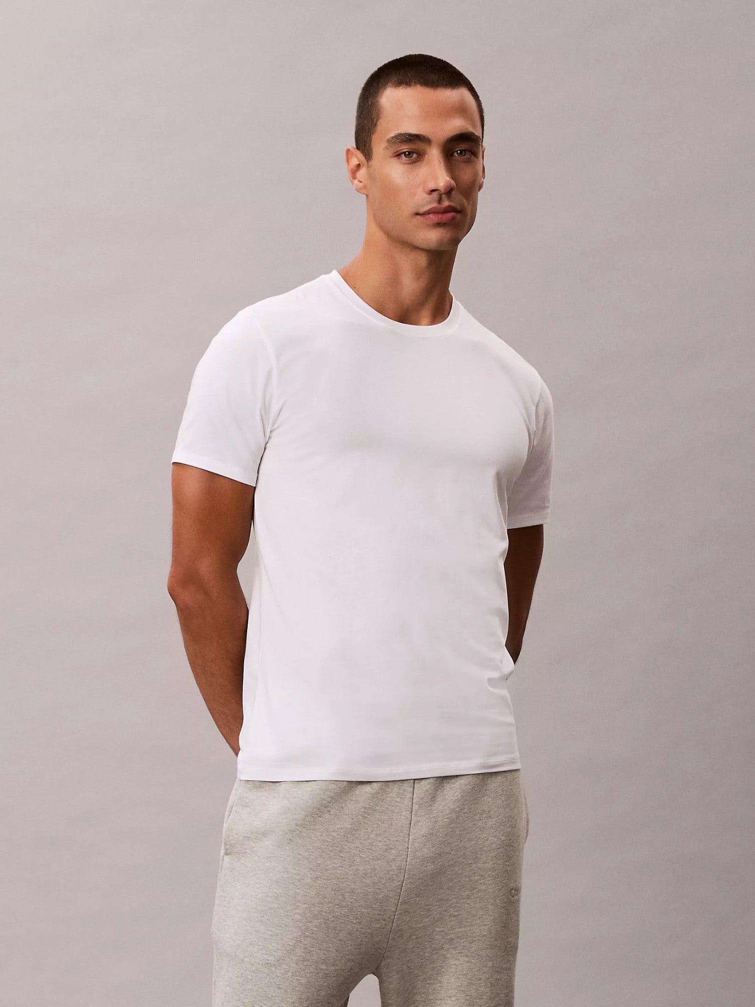 A man wearing a plain white short-sleeve crewneck tshirt and light gray sweatpants, standing against a neutral gray studio background. Calvin Klein, Monza Outlet, Monza Fashion, Shein Outlet