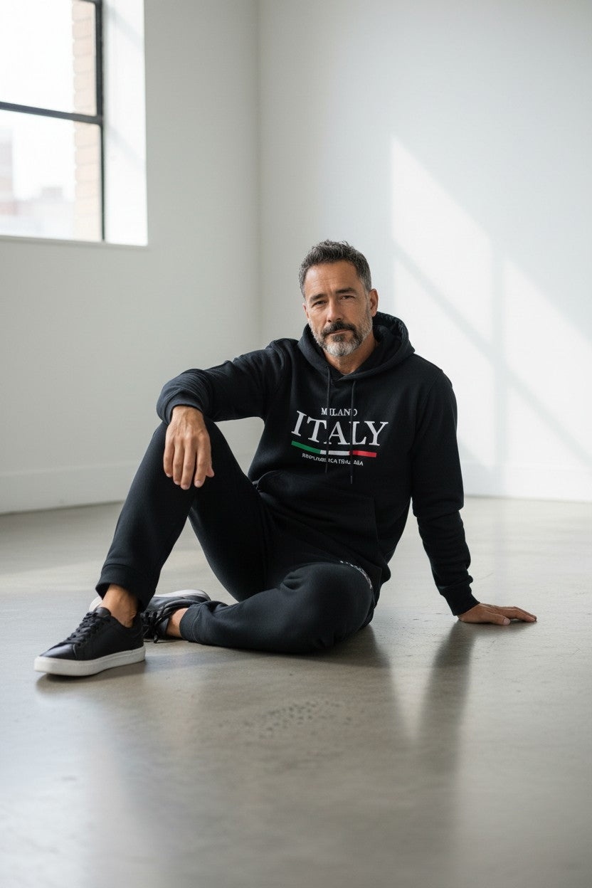 Man sitting on the floor wearing a black hoodie with ITALY text in a modern indoor setting. Antler, Monza Outlet, Monza Fashion, Shein Outlet
