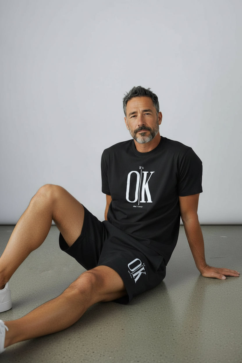 Man wearing a black OK t-shirt and shorts sitting on the floor against a plain background. Antler, Monza Outlet, Monza Fashion, Shein Outlet