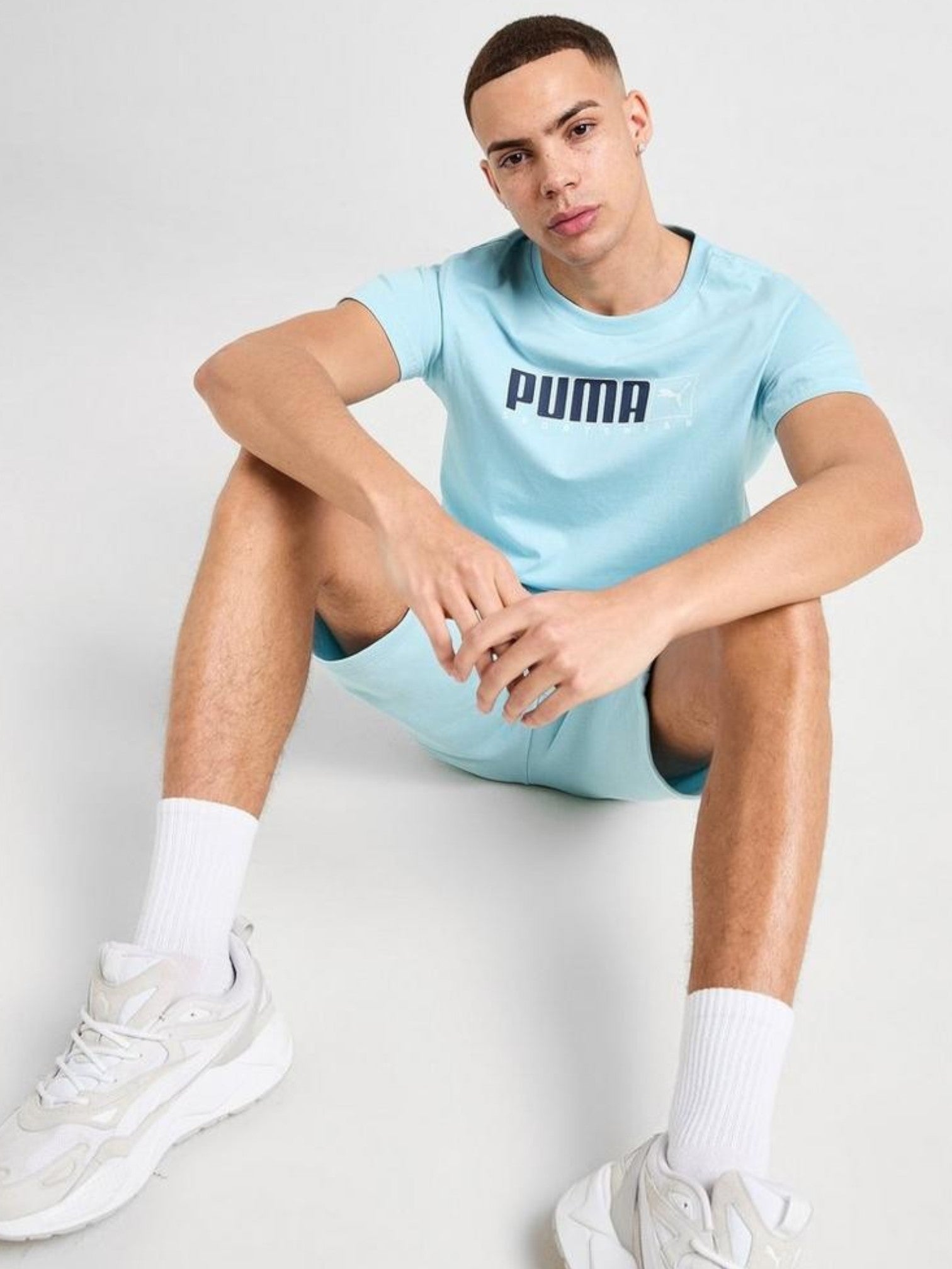 Person wearing a light blue Puma t-shirt and shorts sitting on a white floor. Puma, Monza Outlet, Monza Fashion, Shein Outlet