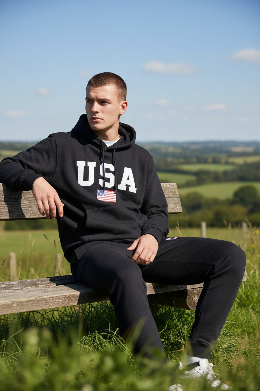 Person wearing a black hoodie with USA and an American flag design, sitting on a wooden bench in a scenic outdoor setting. Antler, Monza Outlet, Monza Fashion, Shein Outlet