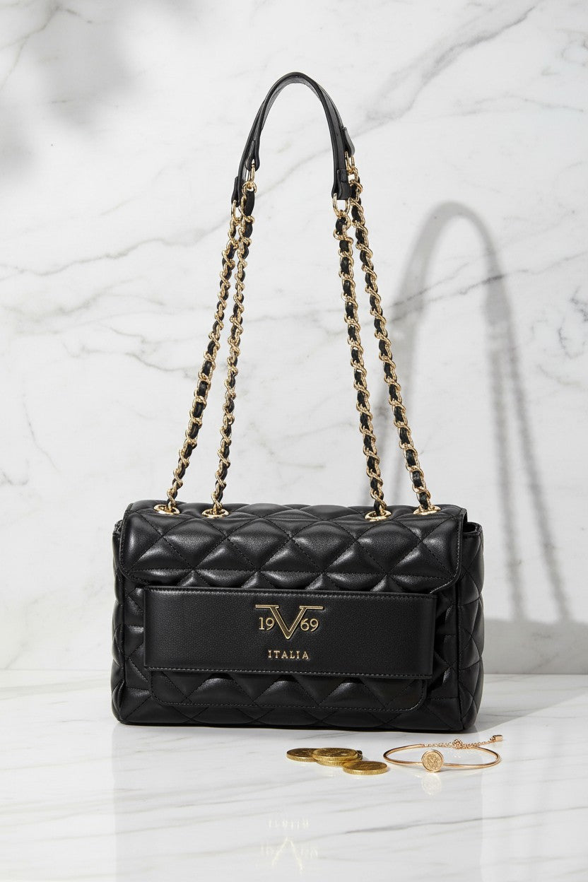 A black quilted leather shoulder bag with gold tone chain straps and a gold tone logo plate on the front. The bag is displayed on a white marble surface with a marble background, accompanied by gold jewelry props. 19V69 Italia, Monza Outlet, Monza Fashion, Shein Outlet