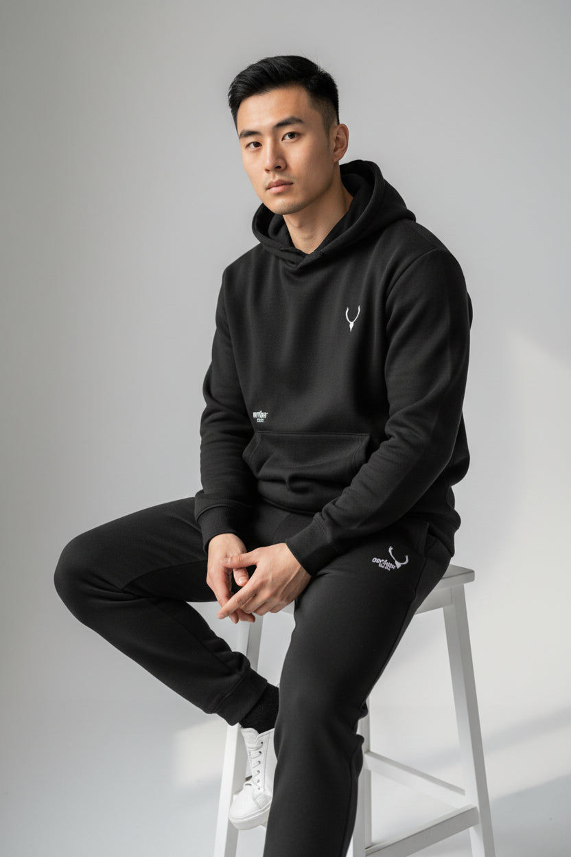 Man wearing a black hoodie and pants sitting on a stool against a gray background. Antler, Monza Outlet, Monza Fashion, Shein Outlet