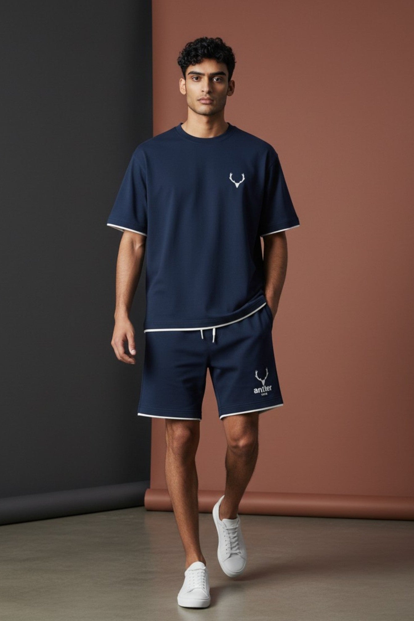 Man wearing navy blue t-shirt and shorts with a logo, standing against a dark and brown gradient wall. Antler Logo Set T-Shirt Shorts Navy Blue, Monza Outlet, Monza Fashion, Shein Outlet.