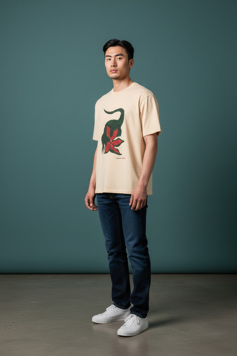 Man wearing a beige t-shirt with a graphic design and dark jeans against a teal background. Lacoste, Monza Outlet, Monza Fashion, Shein Outlet