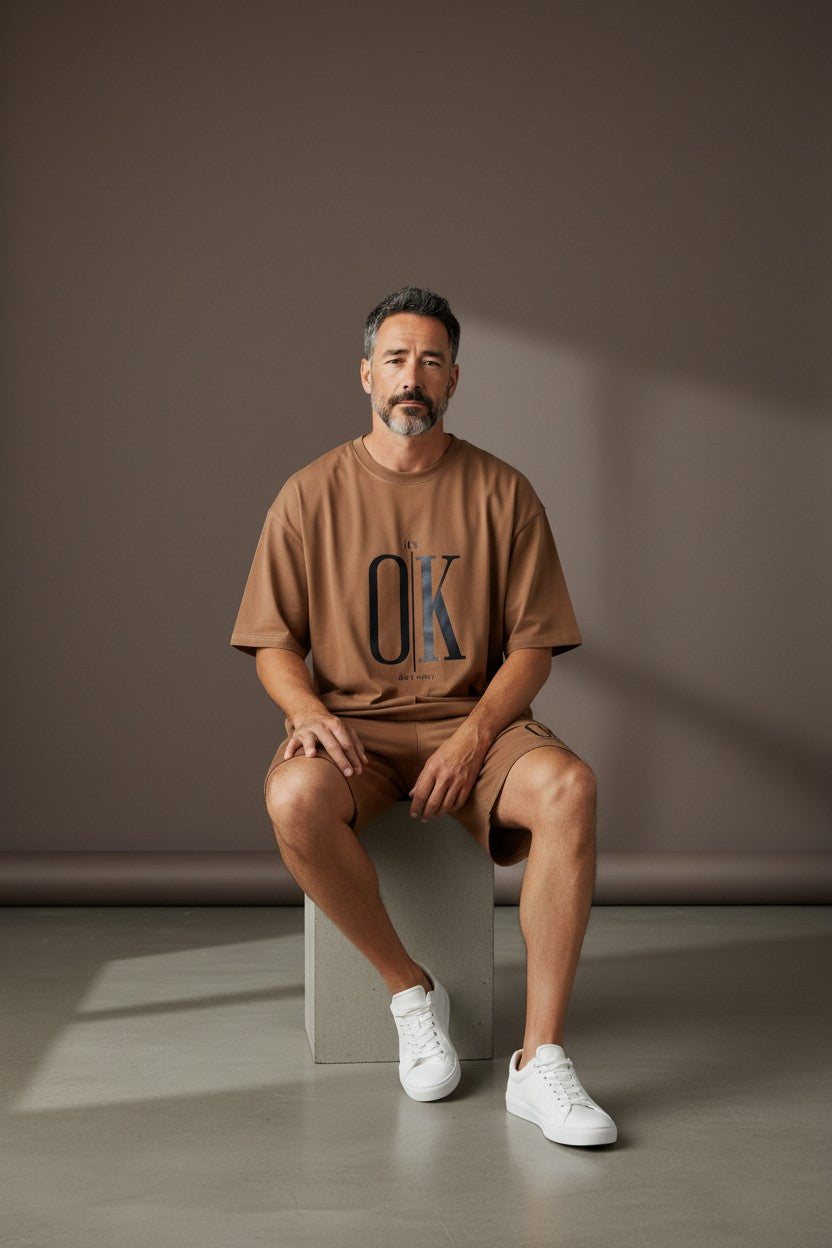 Man sitting on a block wearing a brown OK t-shirt against a plain background. Antler, Monza Outlet, Monza Fashion, Shein Outlet