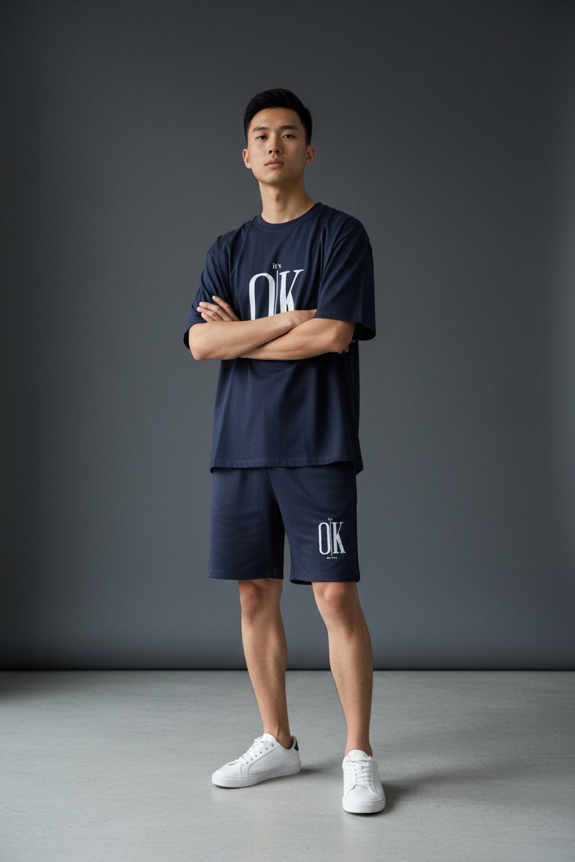 Person wearing navy blue OK branded t-shirt and shorts with white sneakers on a gray background. Antler, Monza Outlet, Monza Fashion, Shein Outlet