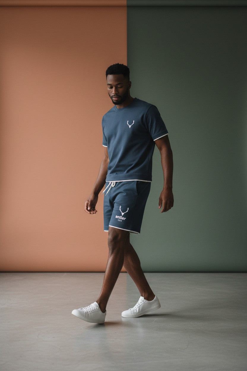 Man wearing a navy blue t-shirt and shorts with a logo, standing against a two-tone wall.