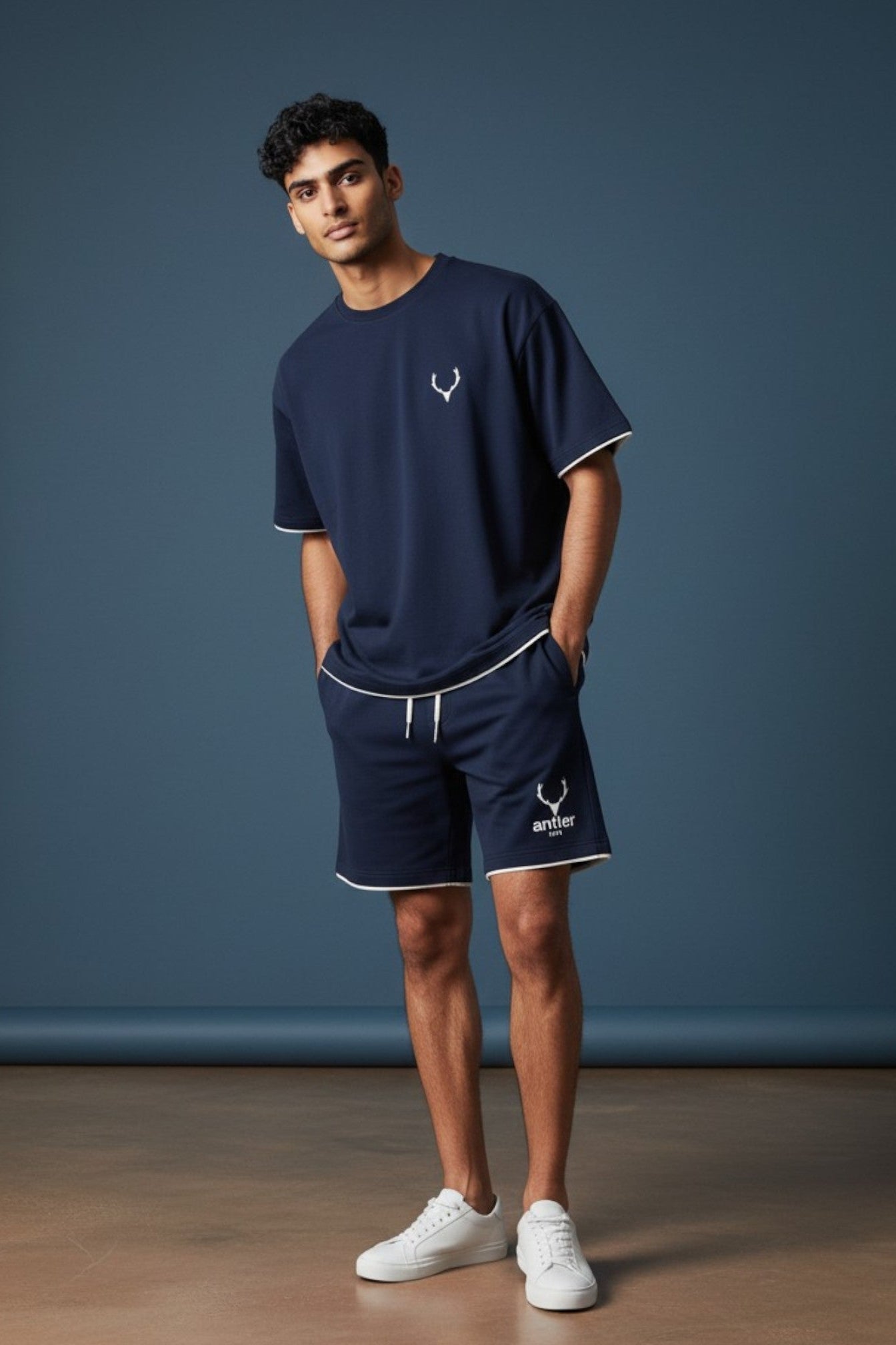 Man wearing navy blue t-shirt and shorts with a logo, standing against a dark blue wall. Antler Logo Set T-Shirt Shorts Navy Blue, Monza Outlet, Monza Fashion, Shein Outlet.