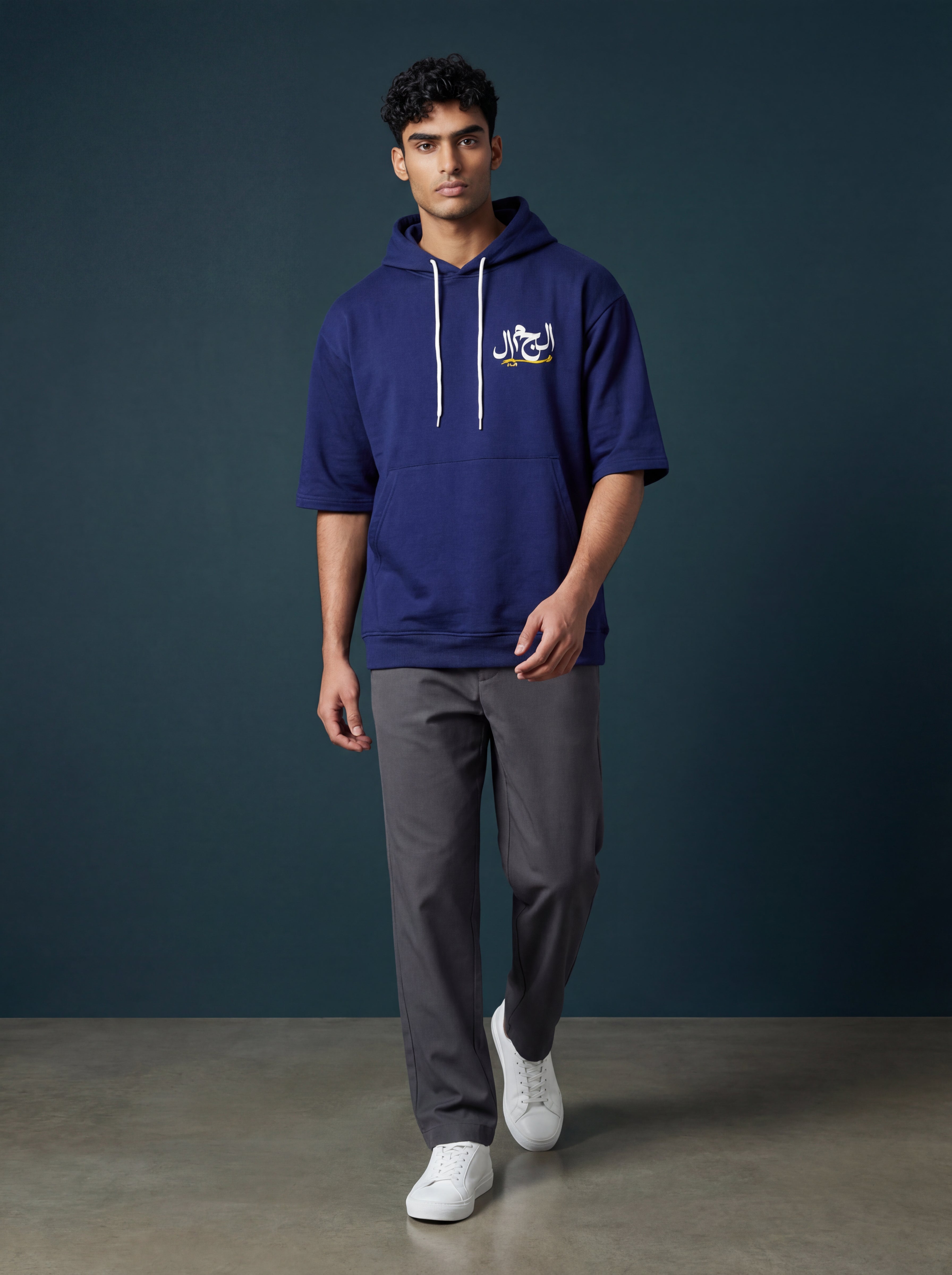 Man wearing a blue hoodie with a logo, gray pants, and white shoes against a dark background. Antler, Monza Outlet, Monza Fashion, Shein Outlet