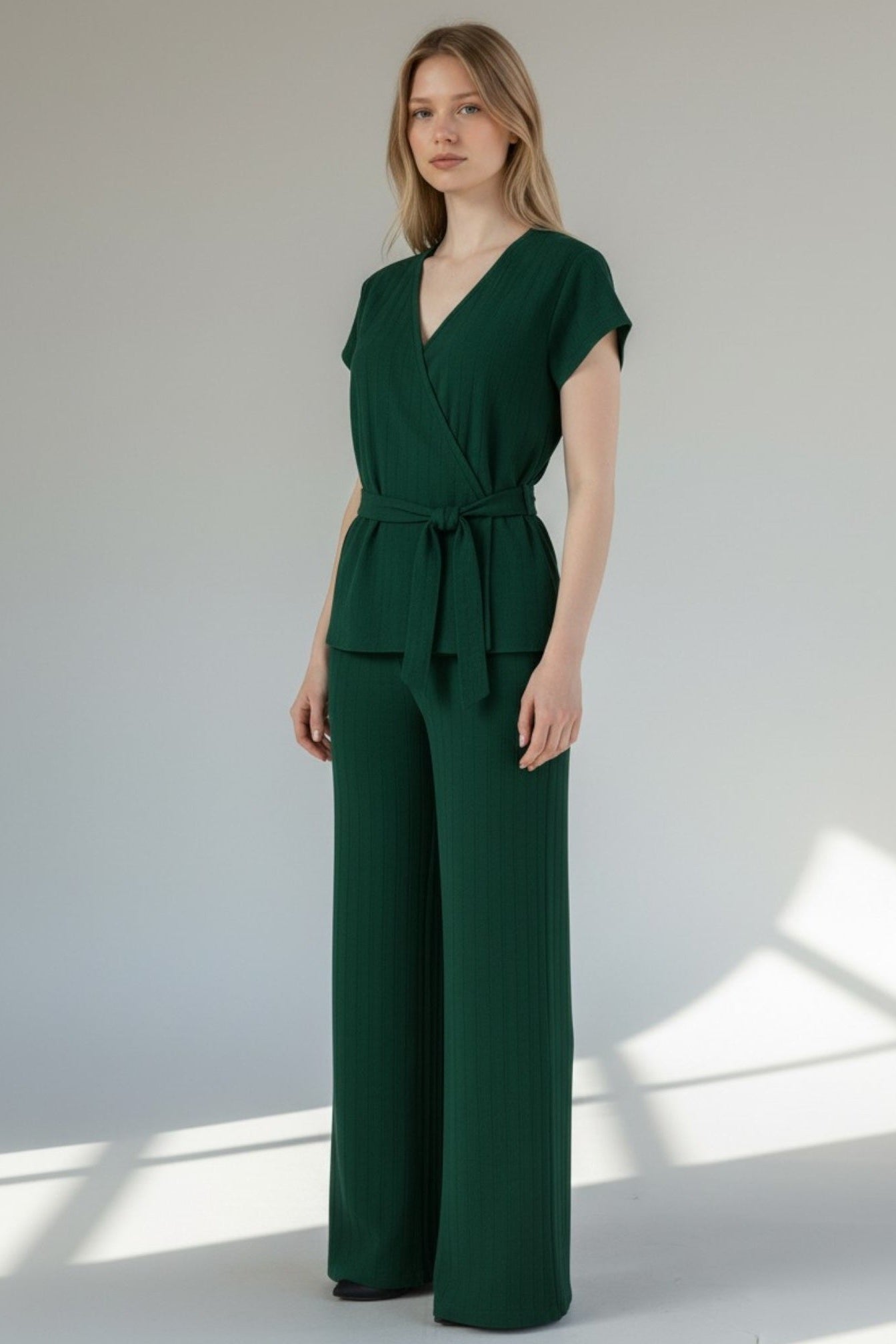 Woman wearing a green jumpsuit standing against a white wall with sunlight casting shadows. Antler, Monza Outlet, Monza Fashion, Shein Outlet