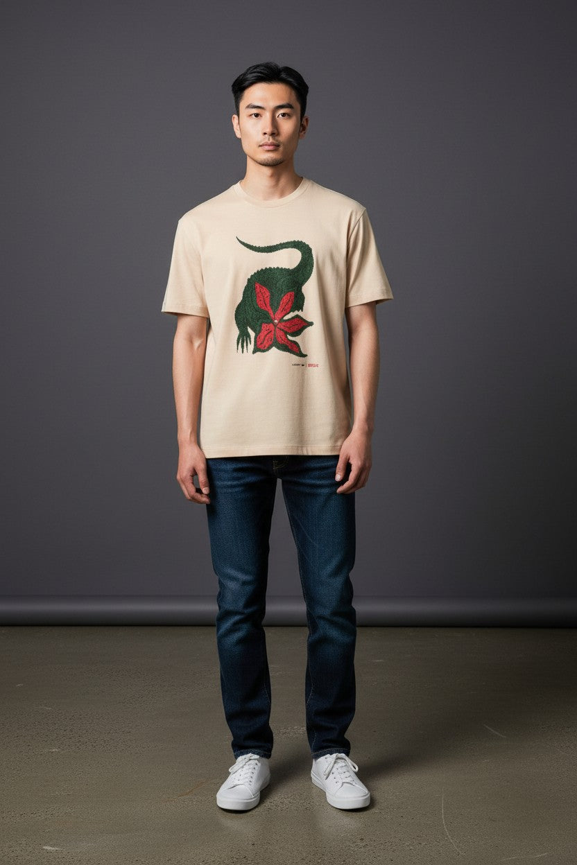 Man wearing a beige t-shirt with a graphic design of a dinosaur and red flower, standing against a plain background. Lacoste, Monza Outlet, Monza Fashion, Shein Outlet