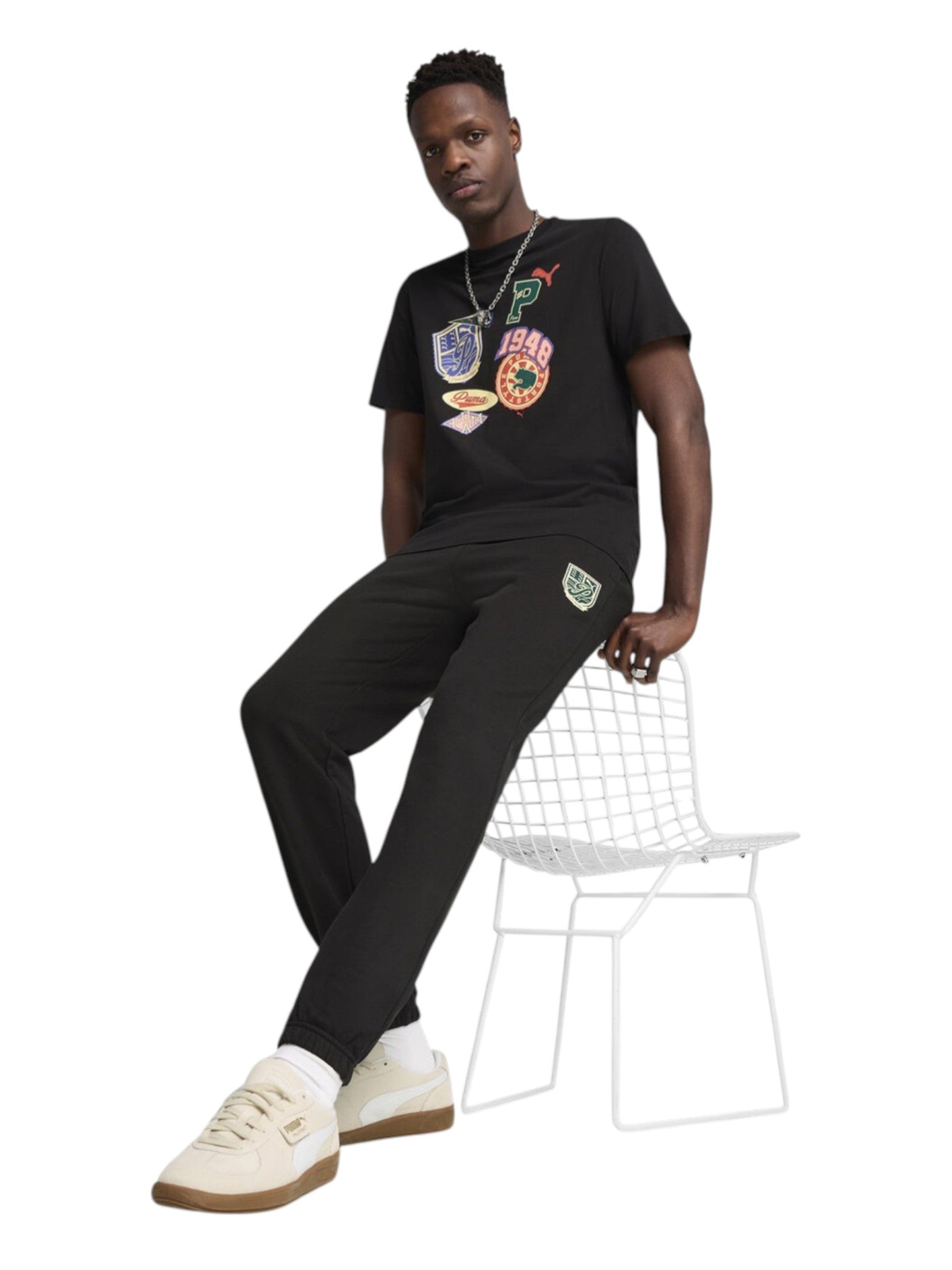 Man wearing a black t-shirt with colorful designs and black pants, sitting on a white chair against a white background. Puma, Monza Outlet, Monza Fashion, Shein Outlet