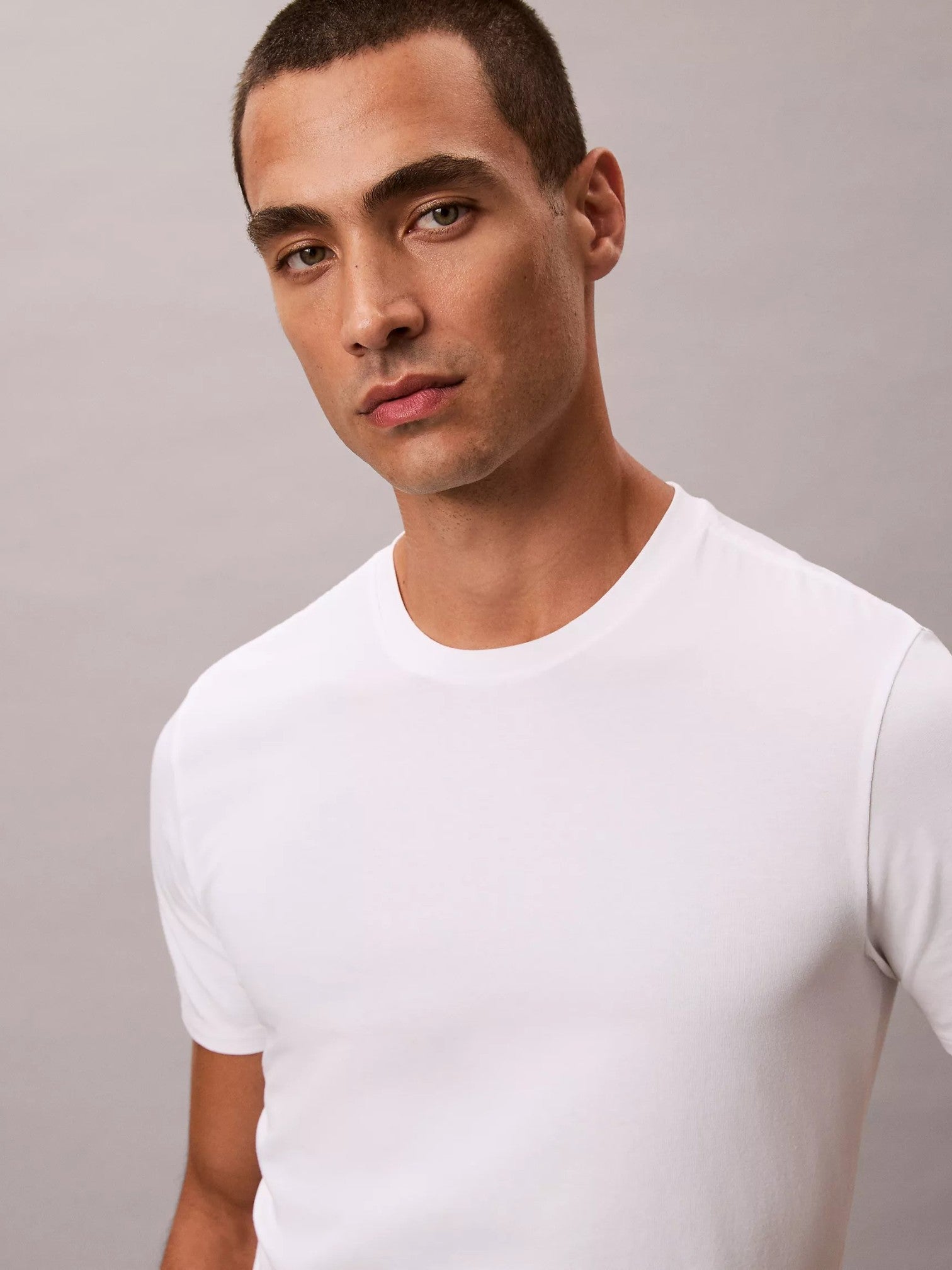 A man wearing a plain white short-sleeve crewneck tshirt, photographed from the chest up against a neutral light gray background. Calvin Klein, Monza Outlet, Monza Fashion, Shein Outlet