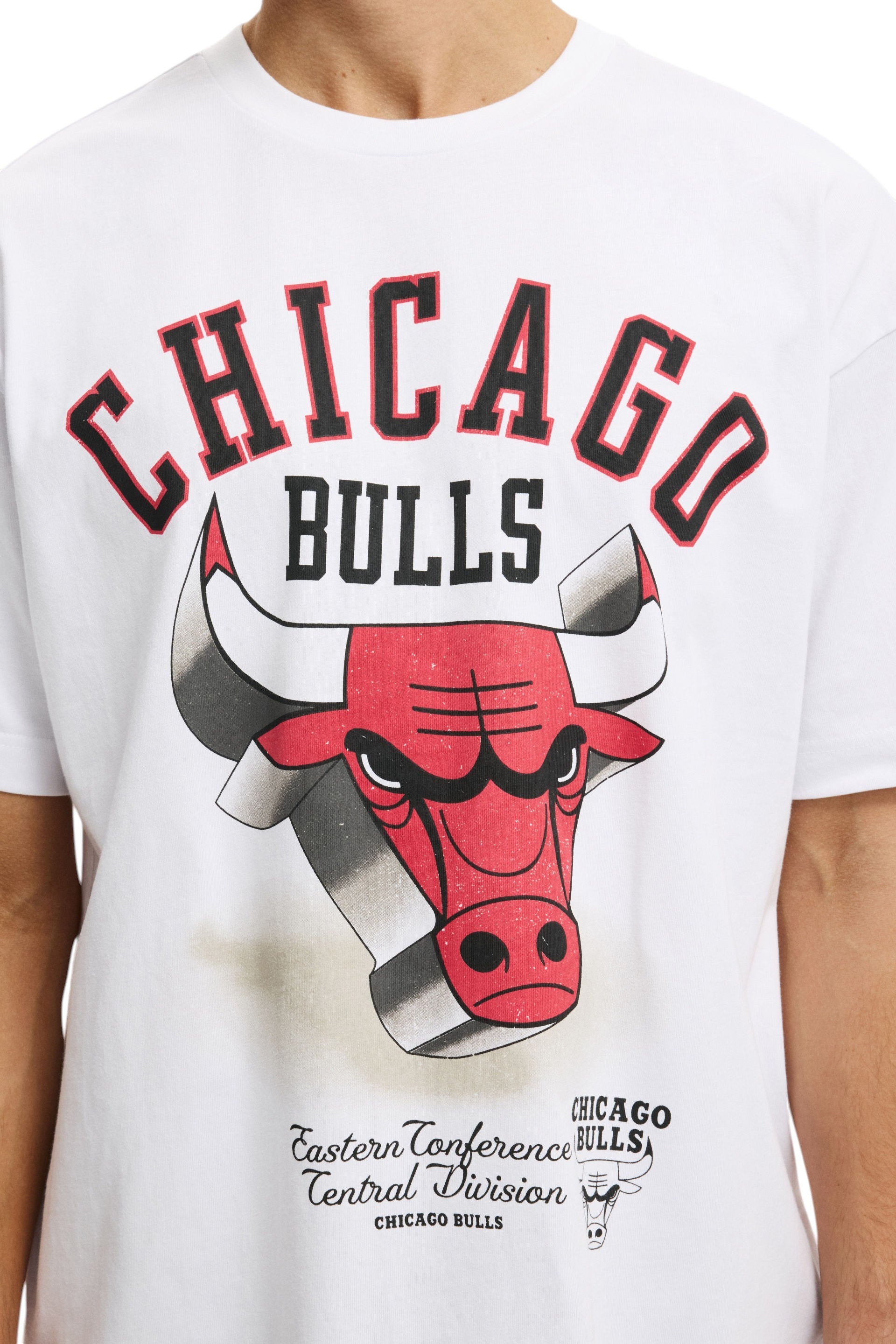 A close-up of a white short-sleeve graphic t-shirt featuring the Chicago Bulls logo and text. The design includes a red bull head with black and gray details, arched CHICAGO text in red and black, and 'BULLS' in black, with Eastern Conference Central Division text below. NBA, Monza Outlet, Monza Fashion, Shein Outlet