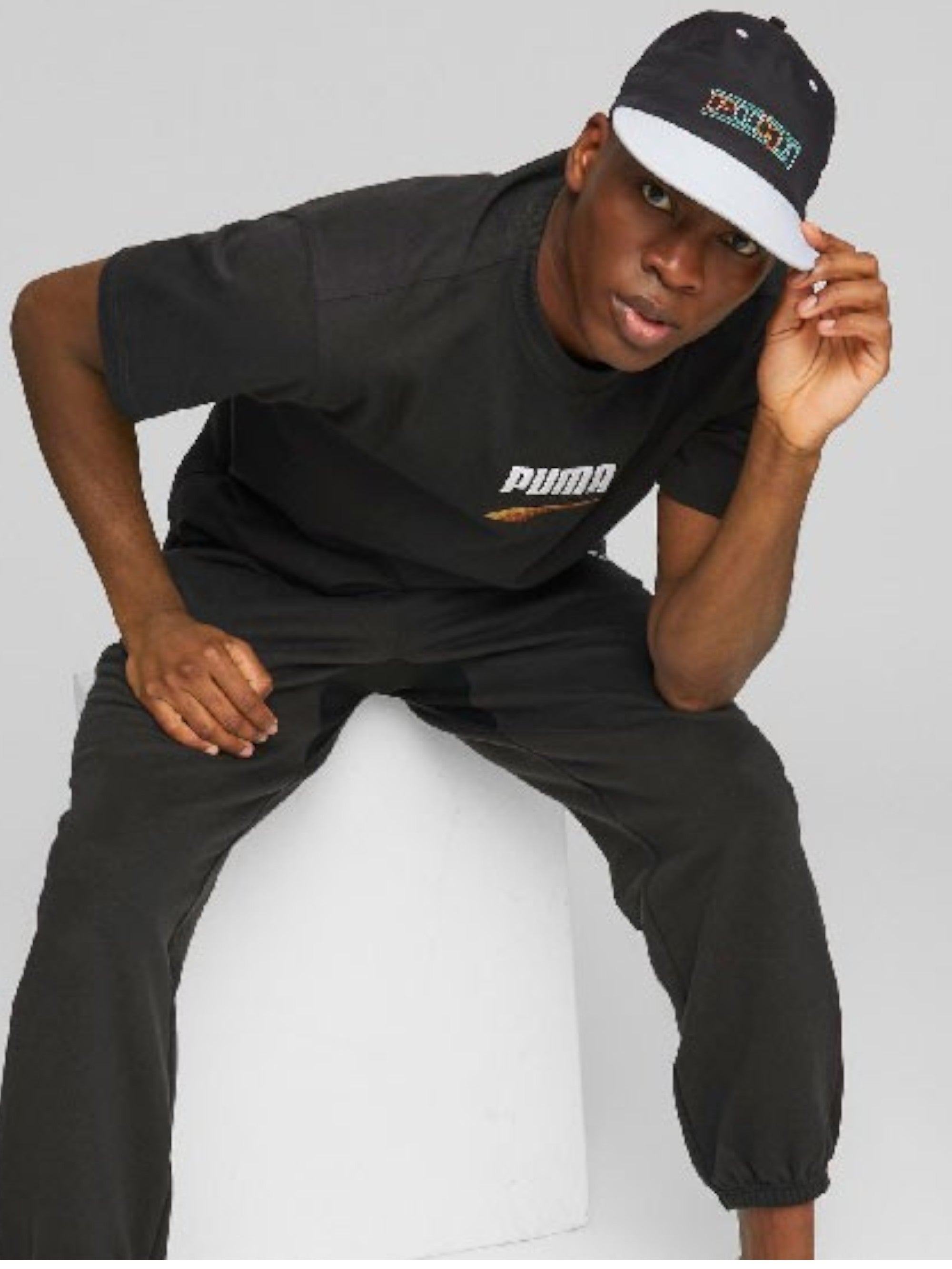 Person wearing a black Puma t-shirt and pants with a cap, sitting on a white stool against a gray background. Puma Monza Outlet