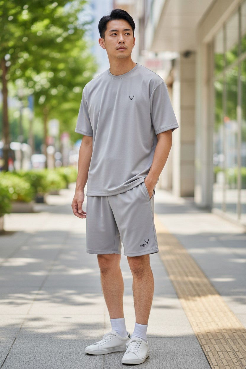 Man wearing a gray t-shirt and shorts standing on a sidewalk with greenery and a building in the background. Antler, Monza Outlet, Monza Fashion, Shein Outlet