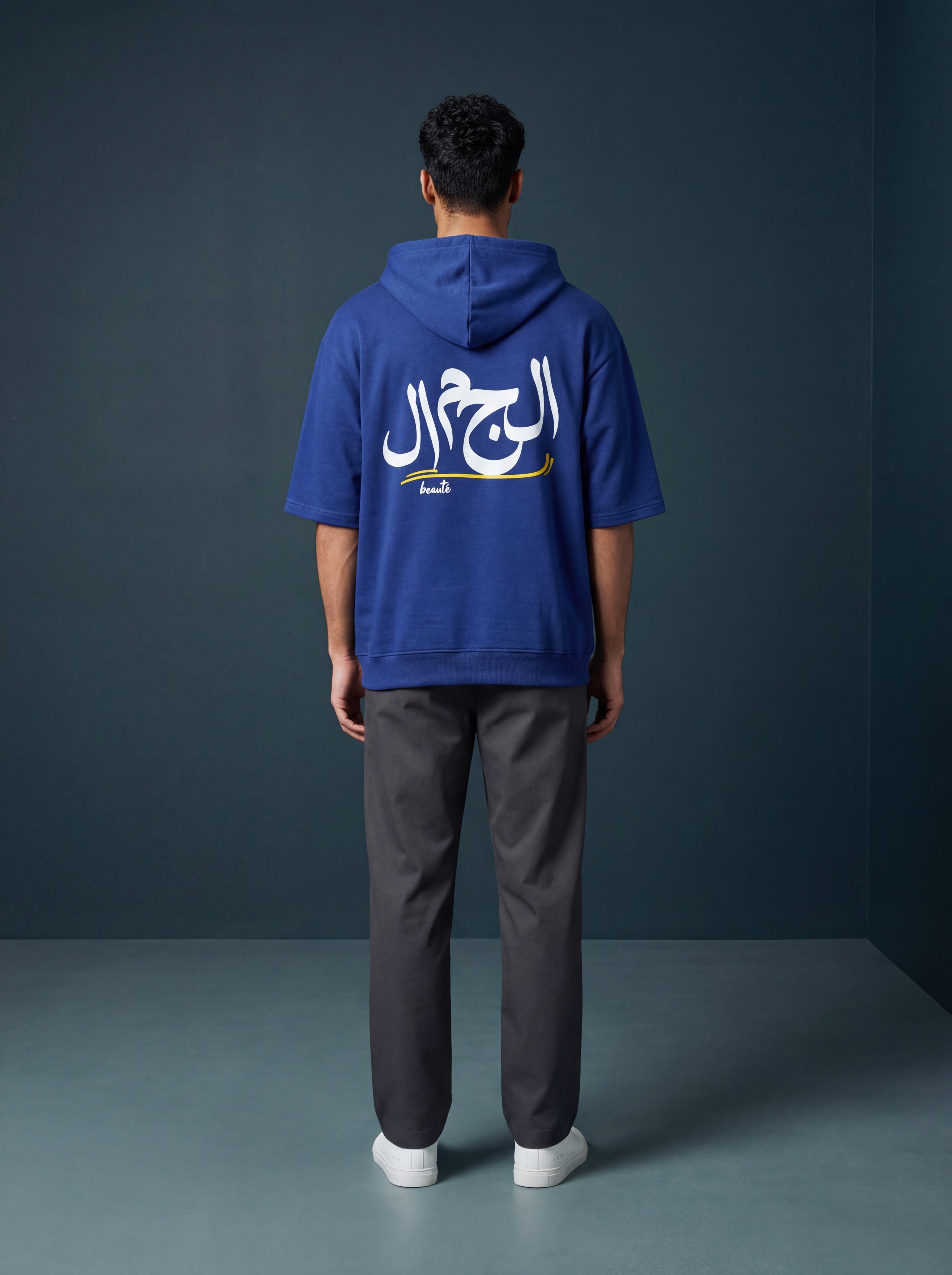 Person wearing a blue hoodie with Arabic text on the back against a dark gray background. Antler, Monza Outlet, Monza Fashion, Shein Outlet  
