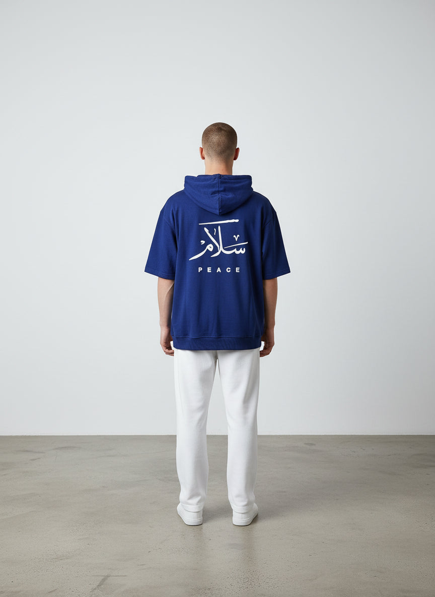 Person wearing a blue hoodie with Arabic text and 'PEACE' on the back, standing against a white wall. Antler, Monza Outlet, Monza Fashion, Shein Outlet  