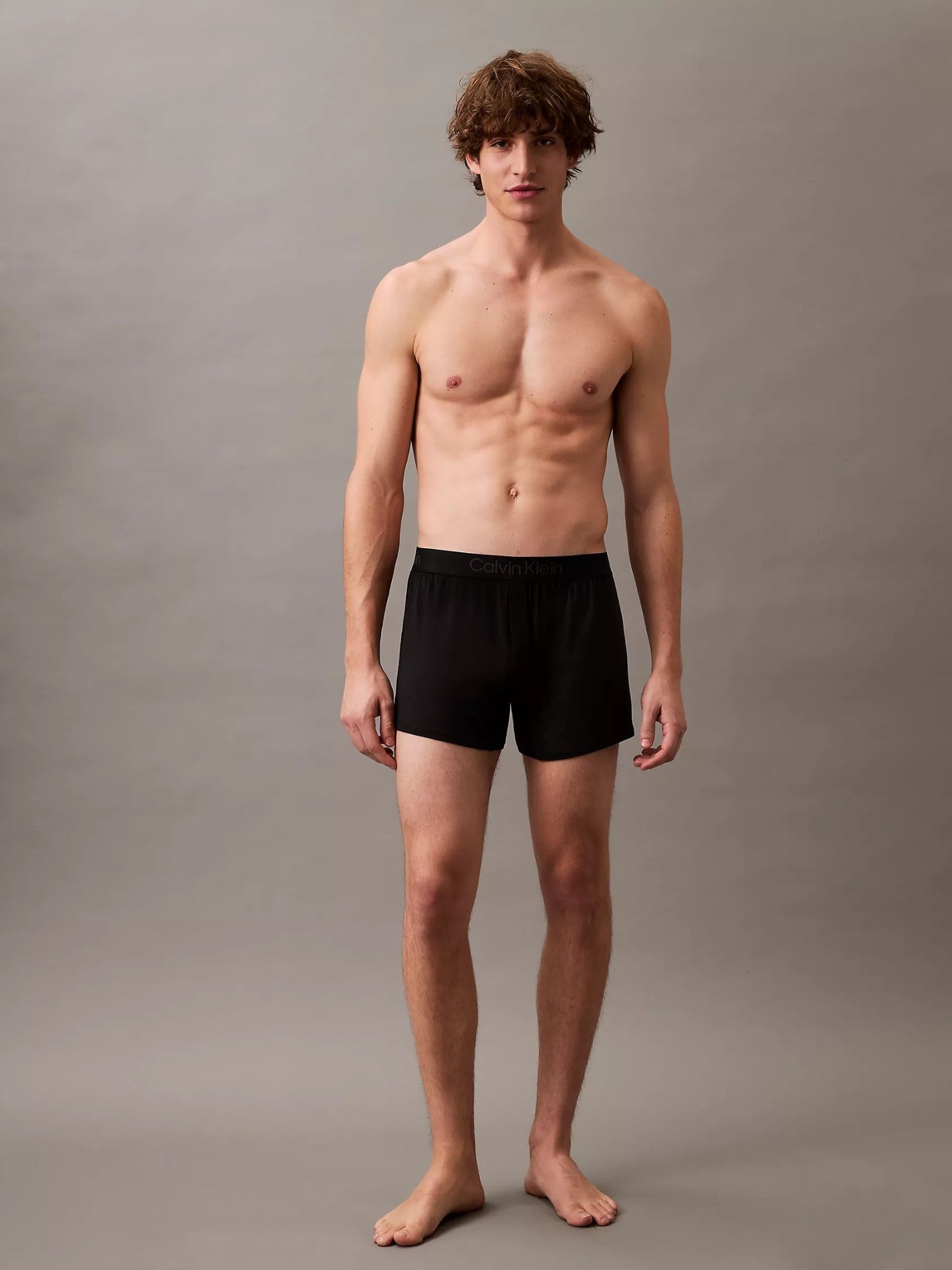 A shirtless male model stands against a plain gray studio background wearing black boxer-brief style shorts. Calvin Klein, Monza Outlet, Monza Fashion, Shein Outlet