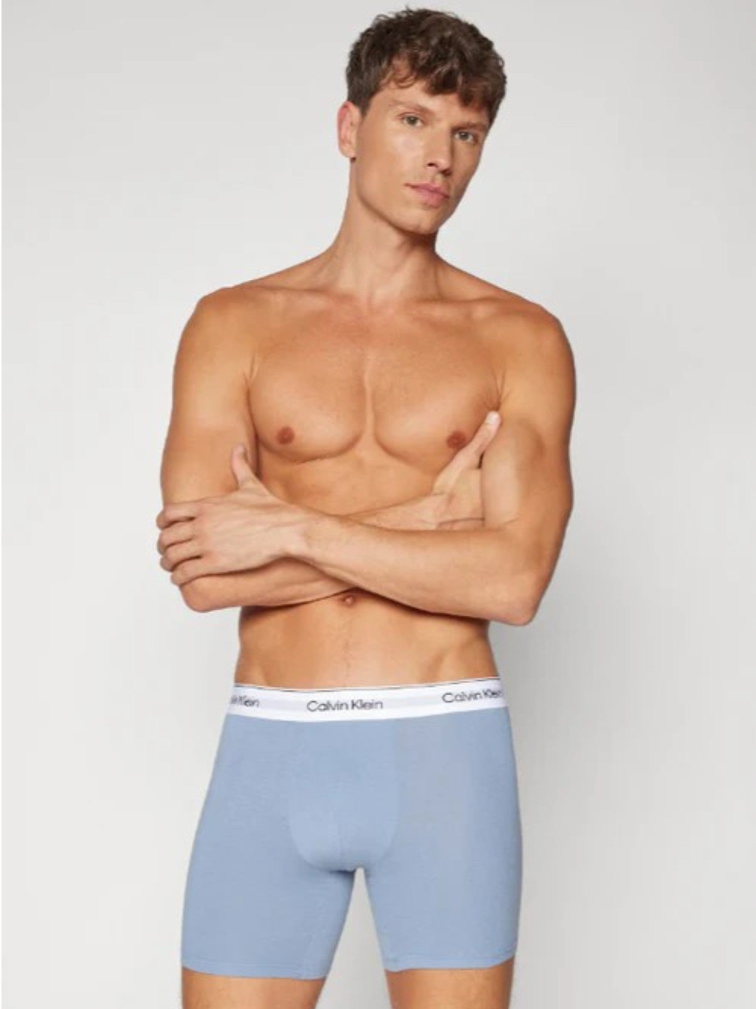 A male model wearing light blue Calvin Klein boxer briefs, standing with arms crossed against a light gray studio background. Calvin Klein, Monza Outlet, Monza Fashion, Shein Outlet