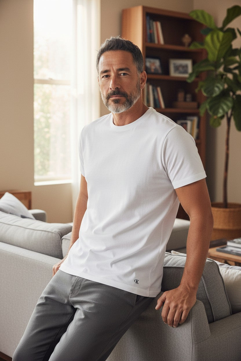 A man wearing a white short-sleeve crewneck t-shirt and gray pants, leaning against a light gray sofa in a modern living room setting. Calvin Klein, Monza Outlet, Monza Fashion, Shein Outlet