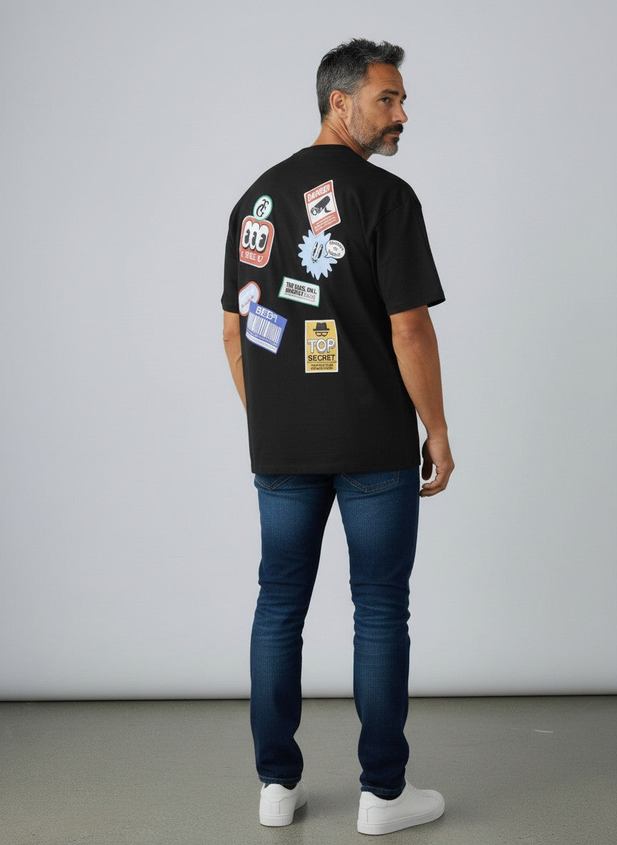 Man wearing a black t-shirt with various stickers on the back, standing against a plain background.  Jack & Jones, Monza Outlet, Monza Fashion, Shein Outlet 