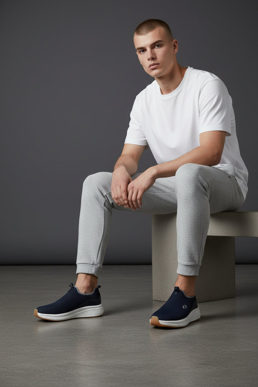 Man sitting on a stool wearing a white t-shirt, gray pants, and navy blue shoes against a dark background. Maklinit, Monza Outlet, Monza Fashion, Shein Outlet