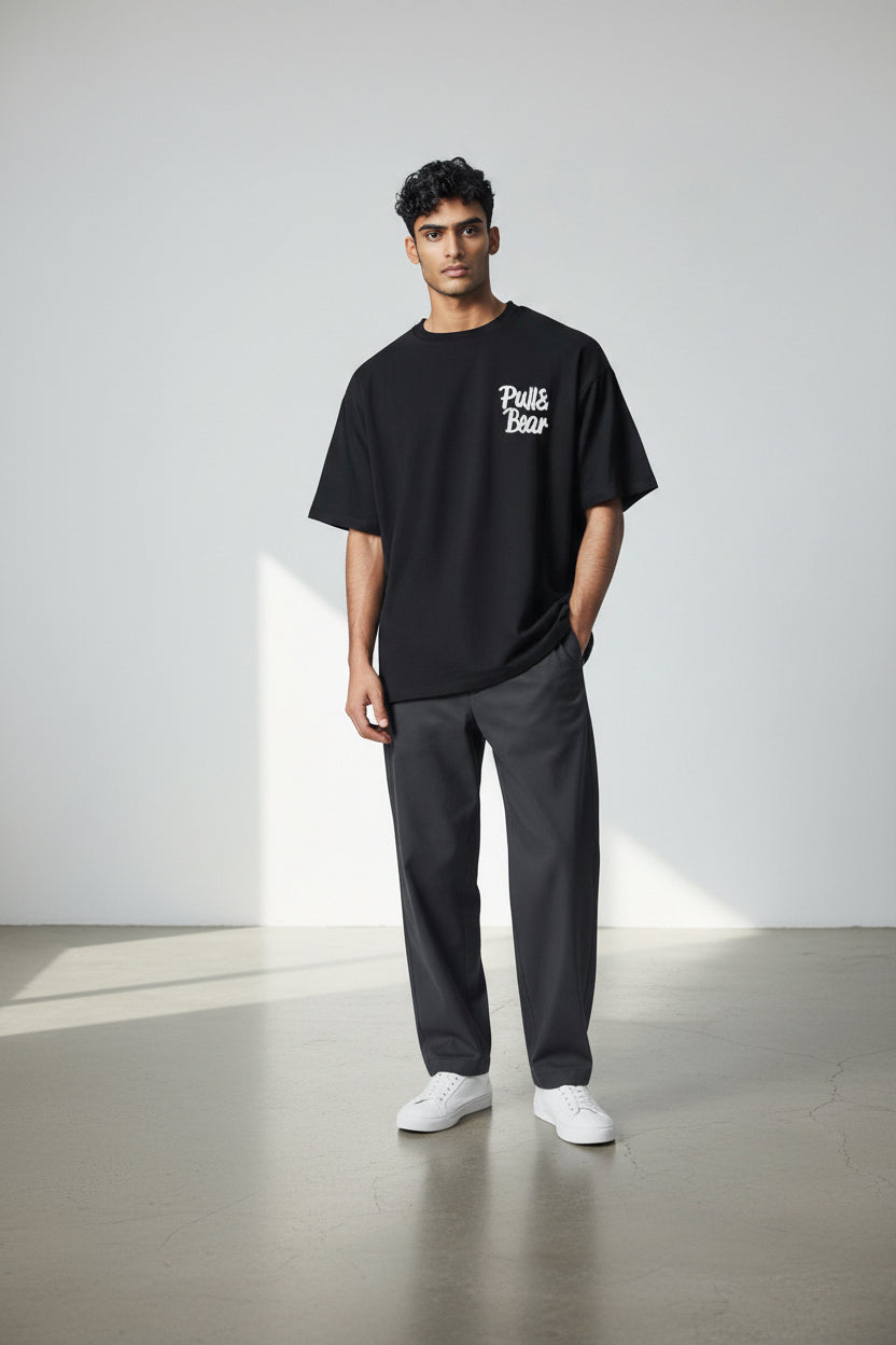 Man wearing a black t-shirt with text, gray pants, and white shoes on a light background. Pull & Bear, Monza Outlet, Monza Fashion, Shein Outlet