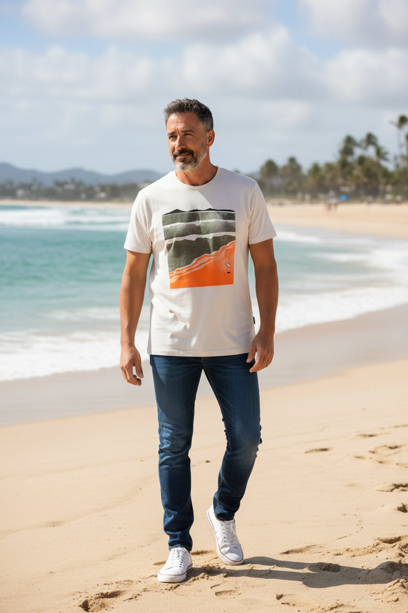 Man standing on a beach wearing a white t-shirt with an orange and black design, blue jeans, and white sneakers. Monza Outlet, Monza Fashion, Shein Outlet