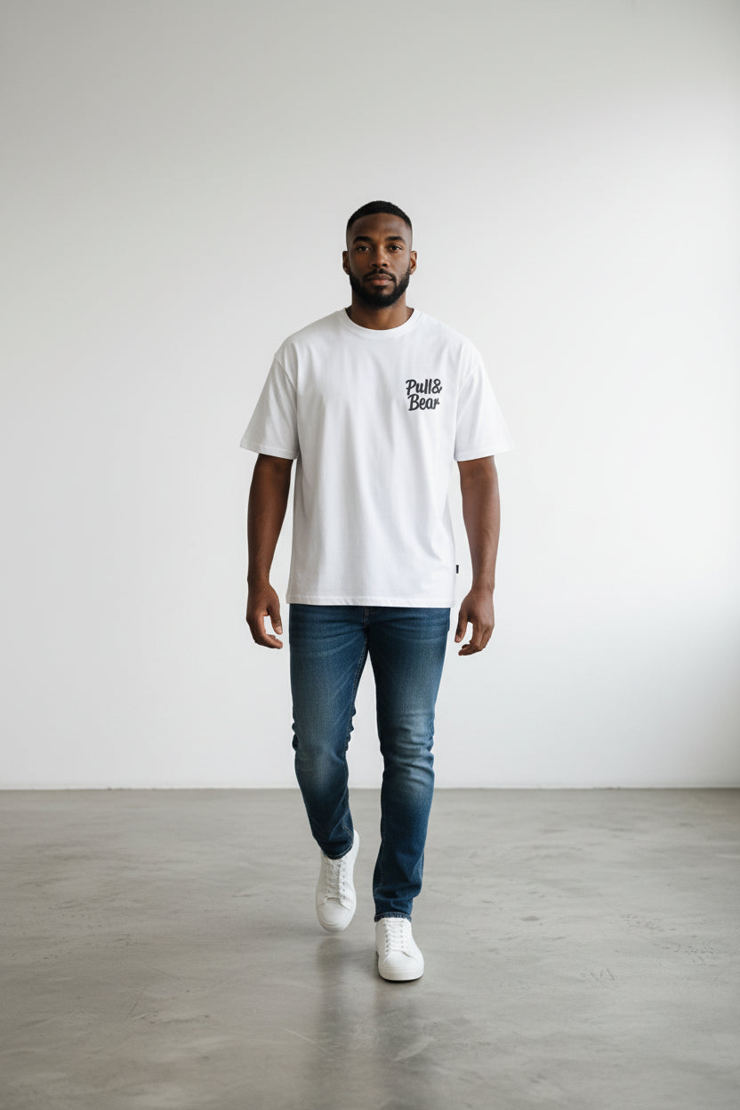 Man wearing a white t-shirt with text and blue jeans standing on a gray floor against a white wall. Pull & Bear, Monza Outlet, Monza Fashion, Shein Outlet