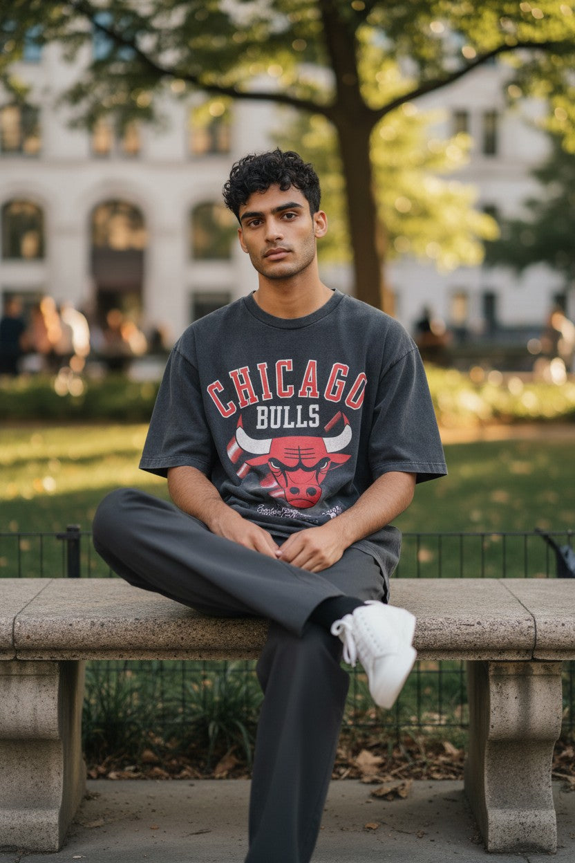 A man sitting on a stone bench in a park wearing a dark gray Chicago Bulls graphic t-shirt, dark gray sweatpants, and white sneakers. NBA, Monza Outlet, Monza Fashion, Shein Outlet