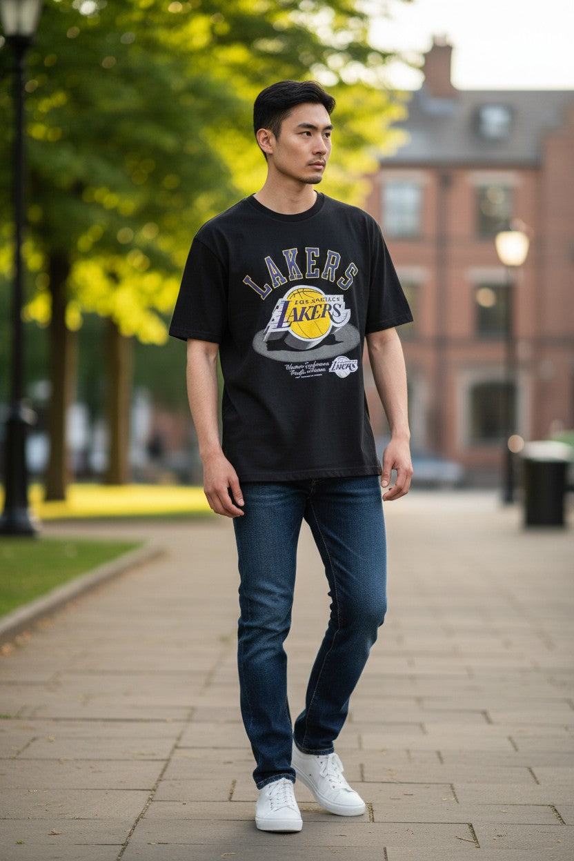 A man wearing a black Los Angeles Lakers graphic t-shirt, blue jeans, and white sneakers walking on a paved outdoor path with trees and a brick building in the background. NBA, Monza Outlet, Monza Fashion, Shein Outlet