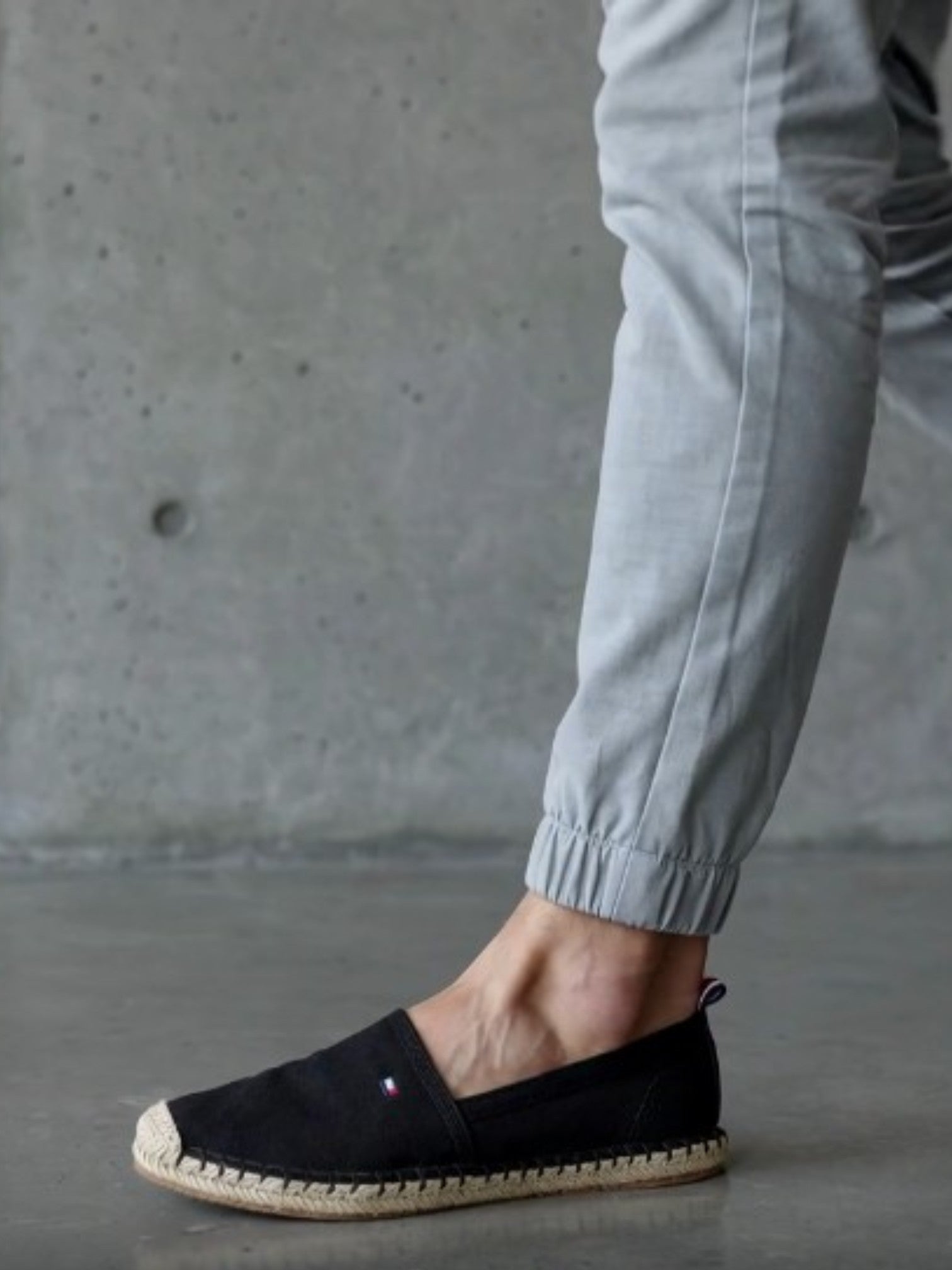 A person wearing a black slip on espadrille shoe with a braided jute sole, paired with light gray pants, standing against a concrete wall. Tommy Hilfiger, Monza Outlet, Monza Fashion, Shein Outlet