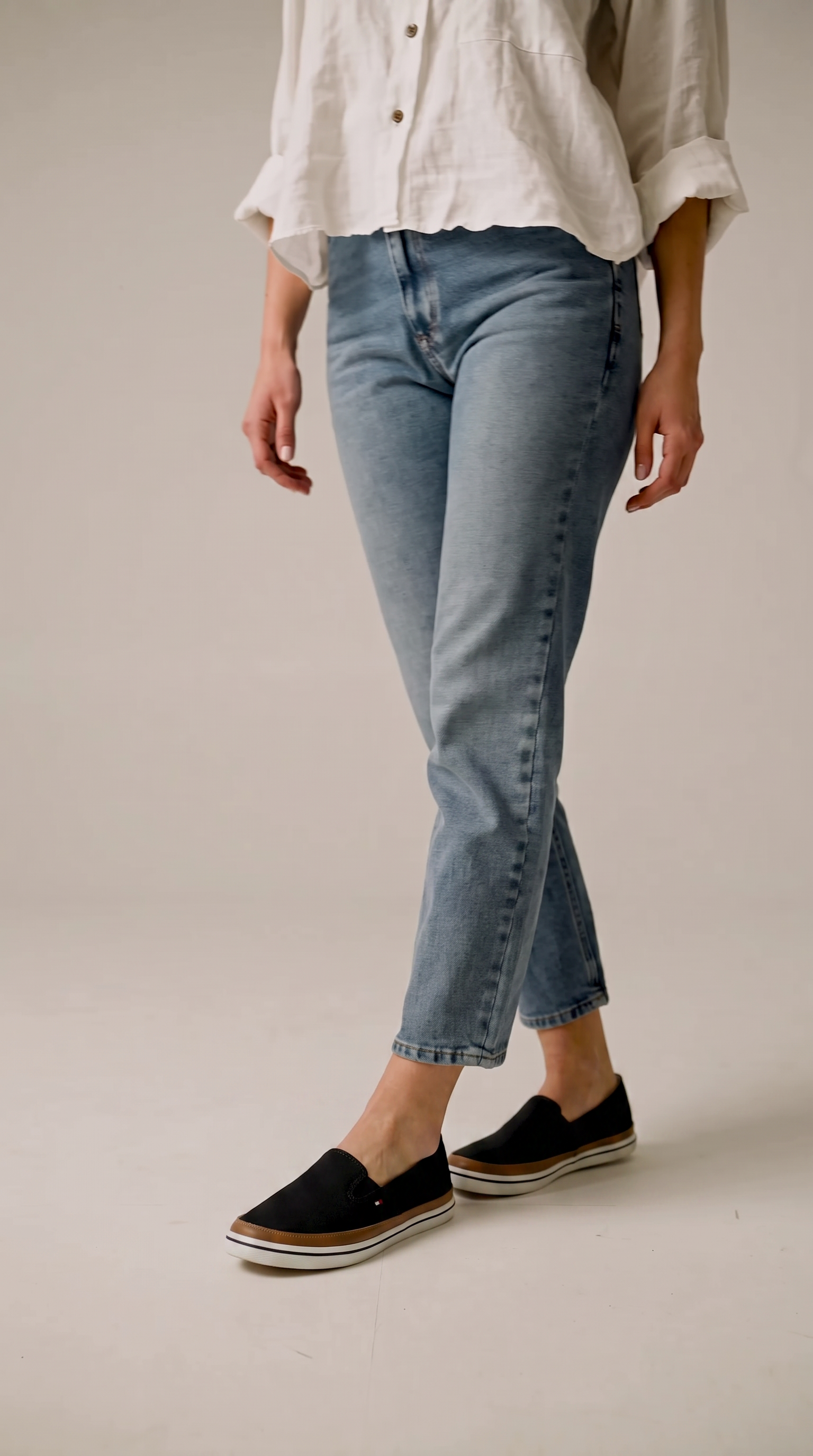 A person wearing light wash blue cropped jeans, a white button-up shirt, and black slip on shoes, standing against a plain light beige studio background. Tommy Hilfiger, Monza Outlet, Monza Fashion, Shein Outlet