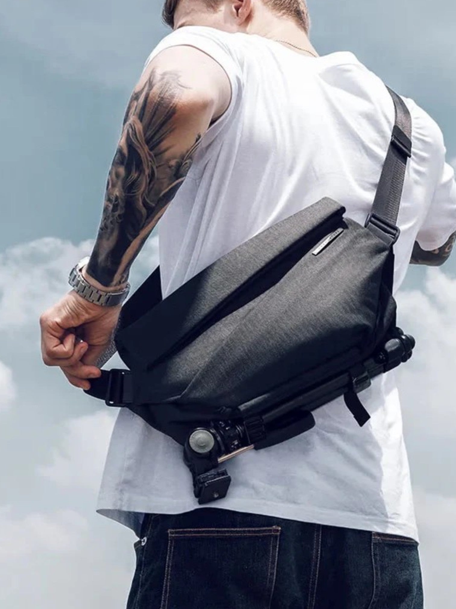 A man wearing a white tank top and dark jeans, viewed from behind, wearing a dark gray crossbody sling bag across his chest against a cloudy sky background. NIIDR, Monza Outlet, Monza Fashion, Shein Outlet
