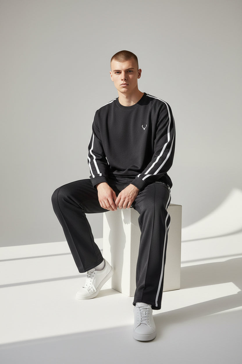 Man wearing a black tracksuit with white stripes sitting on a white stool against a light gray background. Antler, Monza Outlet, Monza Fashion, Shein Outlet