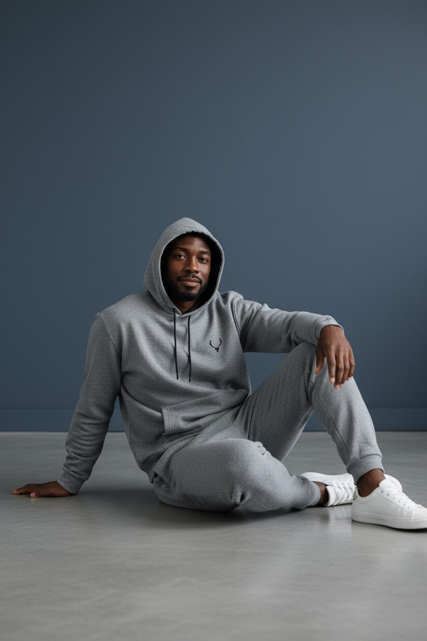 Person wearing a gray hoodie and pants sitting on the floor against a dark blue wall. Antler, Monza Outlet, Monza Fashion, Shein Outlet