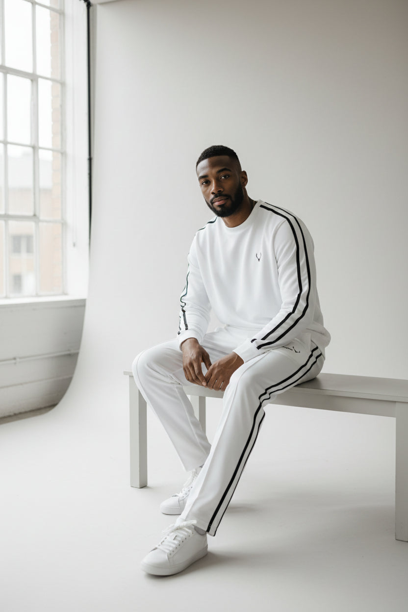 Man wearing a white tracksuit with black stripes sitting on a white bench in a minimalistic room. Antler, Monza Outlet, Monza Fashion, Shein Outlet