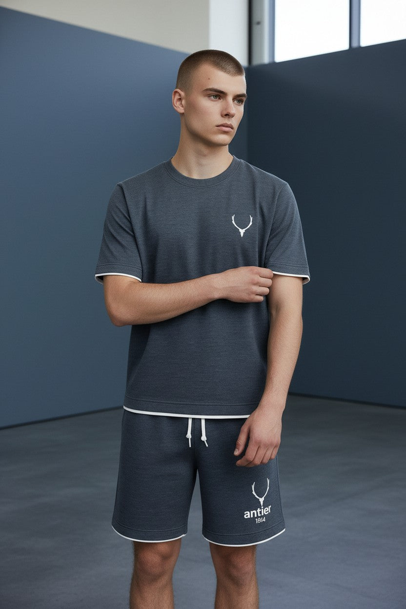 Man wearing a dark gray t-shirt and shorts with a logo, standing against a blue wall. Antler, Monza Outlet, Monza Fashion, Shein Outlet
