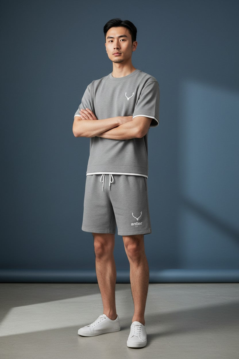 Man wearing a gray t-shirt and shorts set with a logo, standing against a blue wall. Antler, Monza Outlet, Monza Fashion, Shein Outlet