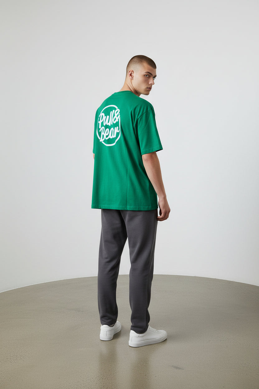 Person wearing a green t-shirt with a logo on the back, standing against a plain white background. Pull & Bear, Monza Outlet, Monza Fashion, Shein Outlet