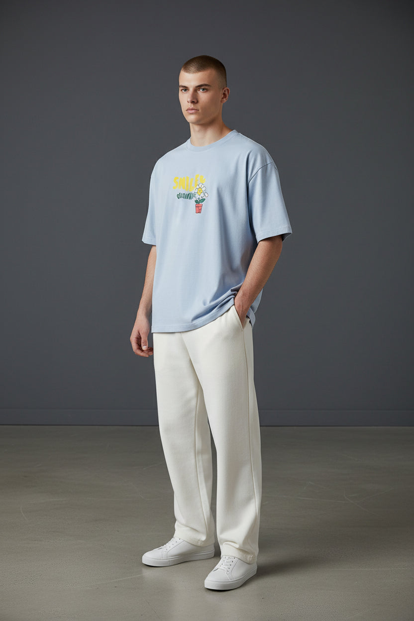 Man wearing a light blue t-shirt with colorful graphics and white pants against a gray background. Monza Outlet, Monza Fashion, Shein Outlet
