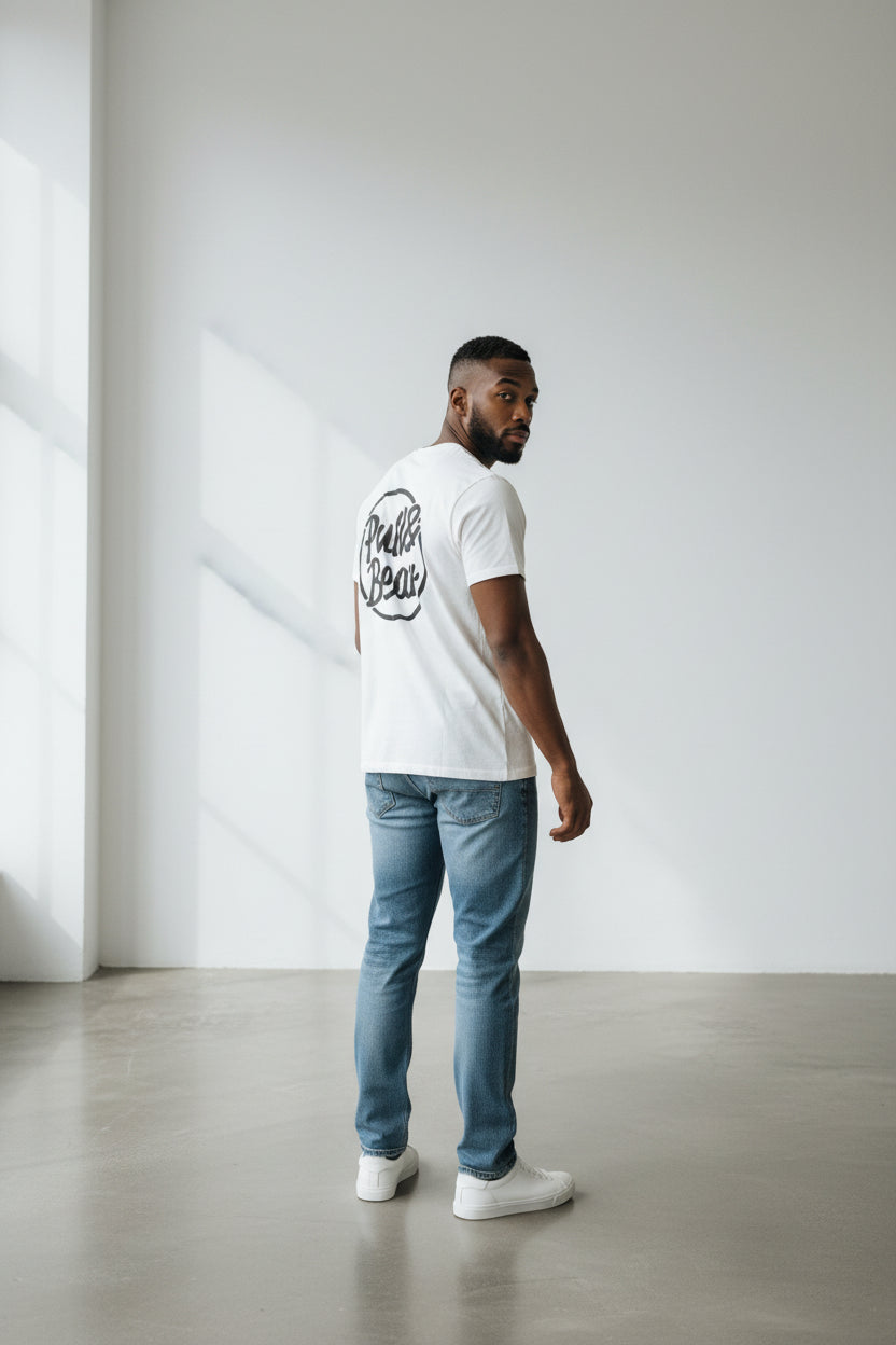 Man wearing a white t-shirt with text and light blue jeans standing against a plain white wall. Pull & Bear, Monza Outlet, Monza Fashion, Shein Outlet