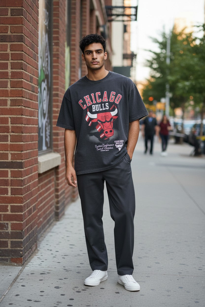 A man standing on a city sidewalk wearing a dark gray Chicago Bulls graphic t-shirt, dark gray sweatpants, and white sneakers, with a brick building in the background. NBA, Monza Outlet, Monza Fashion, Shein Outlet