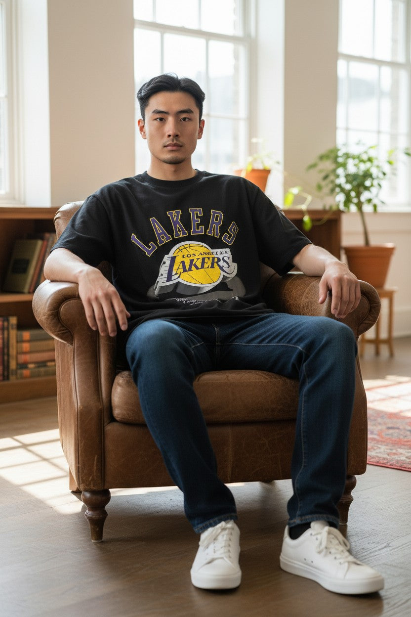 A man sits in a brown leather armchair wearing a black Los Angeles Lakers graphic t-shirt, dark blue jeans, and white sneakers in a bright, modern room with large windows. NBA, Monza Outlet, Monza Fashion, Shein Outlet